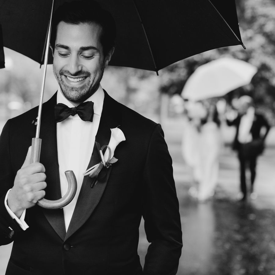 Groom in his tux walks with an umbrella on his way to see the bride at his wedding