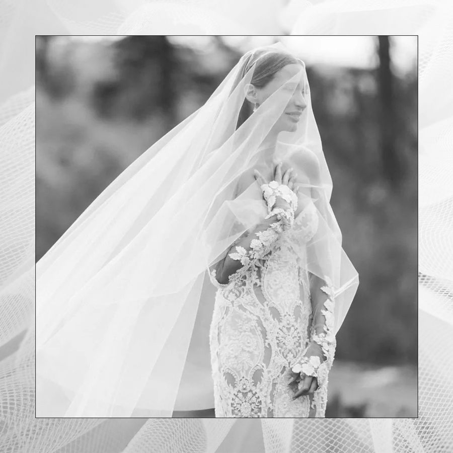 A bride walking, wearing a lace white wedding dress with a long lace veil draped over her face.