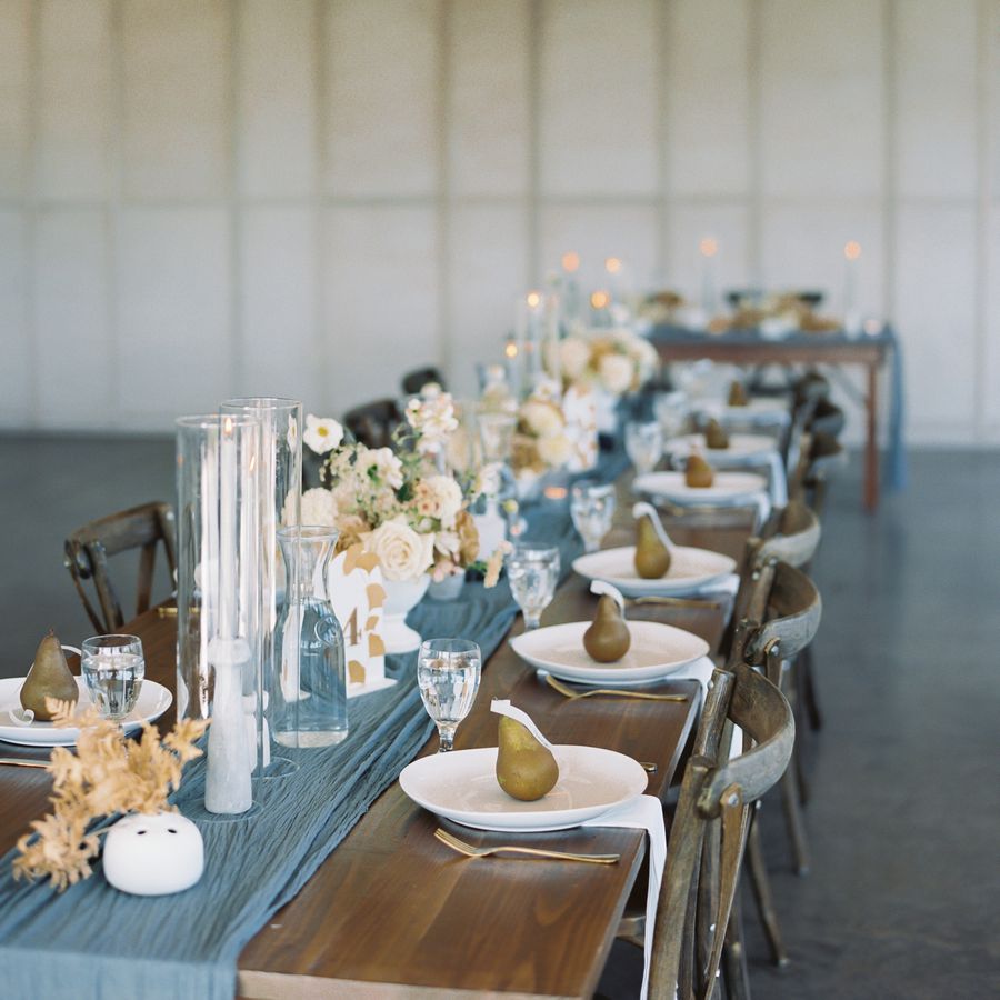 A rehearsal dinner table set with a blue runner, tall candles, white roses, and pear-shaped candle guest favors. 