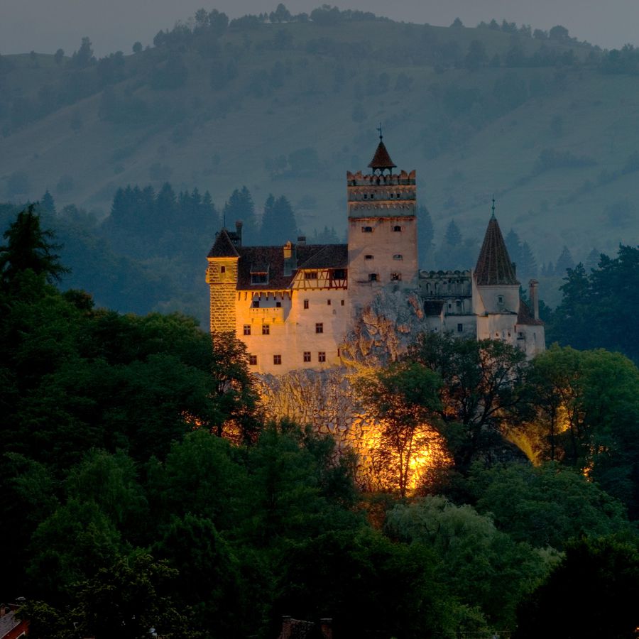 A spooky stone castle with turrets lit up at dusk, surrounded by trees and hills.