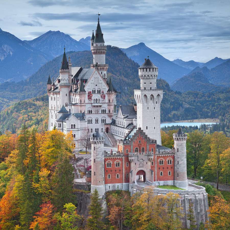 An aerial view of a castle with turrets set amongst fall foliage with mountains in the background.