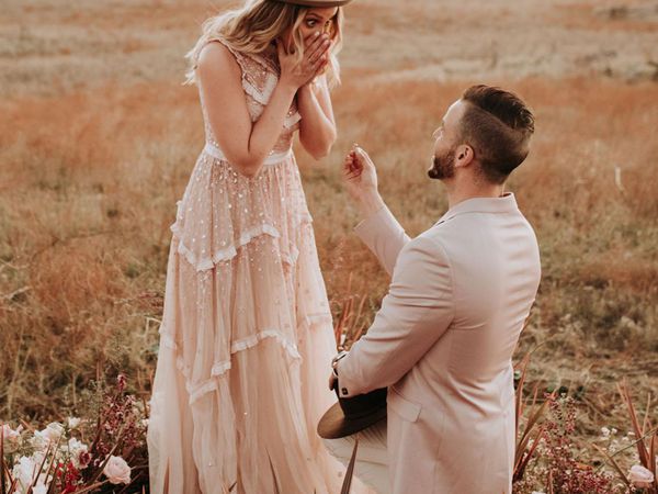 Man Down on One Knee Proposing to Woman in Long Gown and Hat Outside