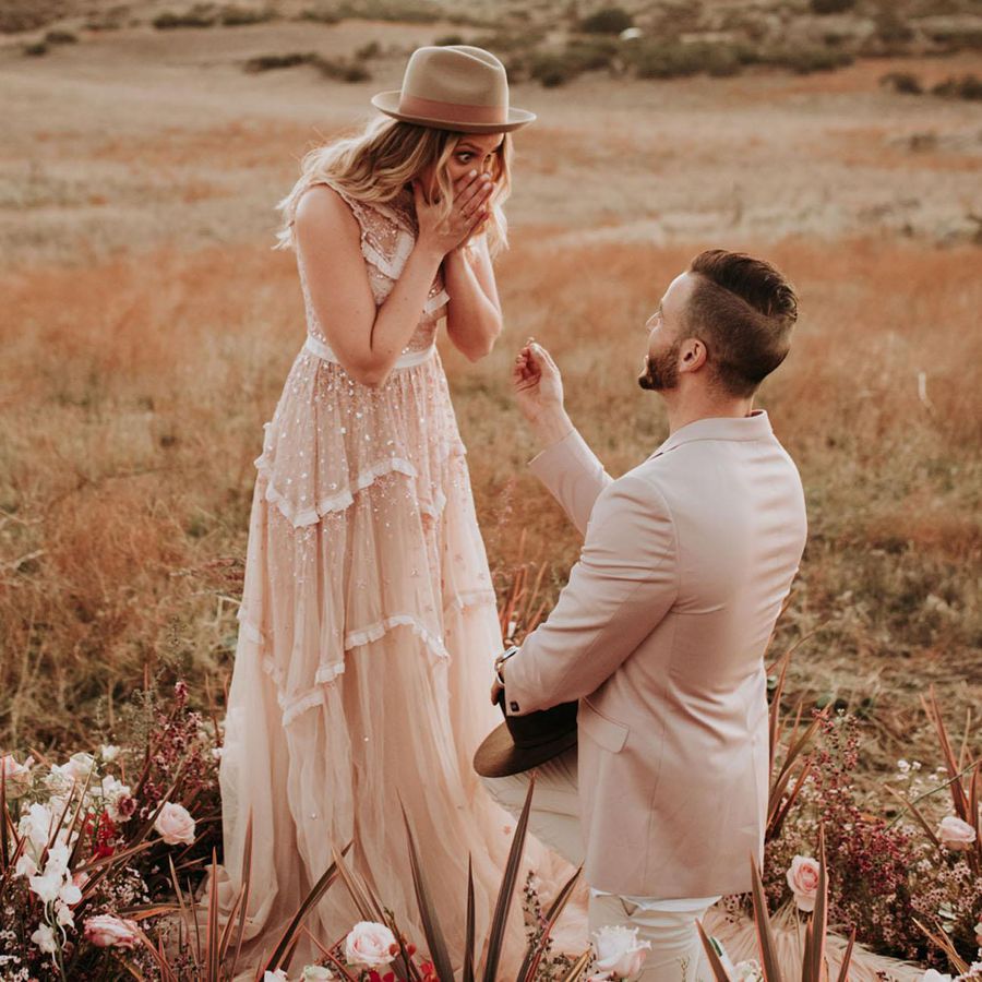 Man Down on One Knee Proposing to Woman in Long Gown and Hat Outside