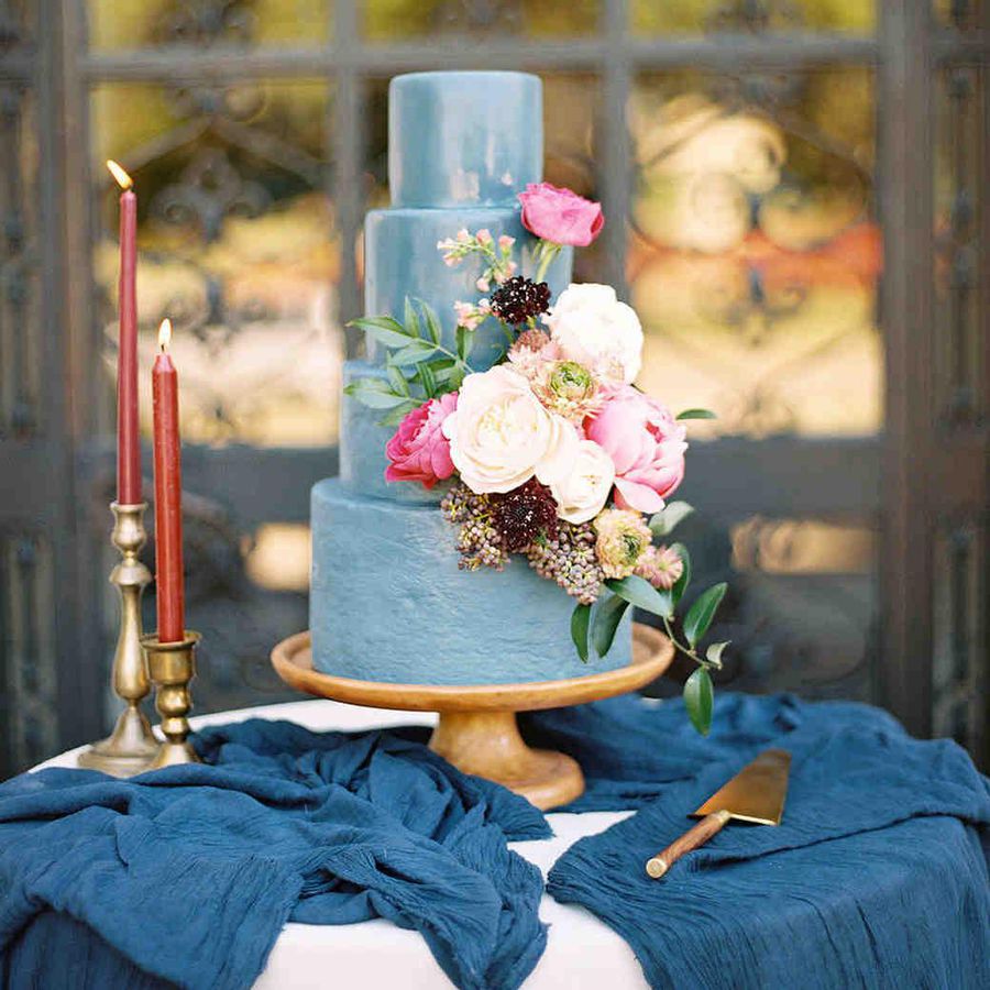 Four-tier blue wedding cake with pink, white, and dark florals on cake stand and blue table