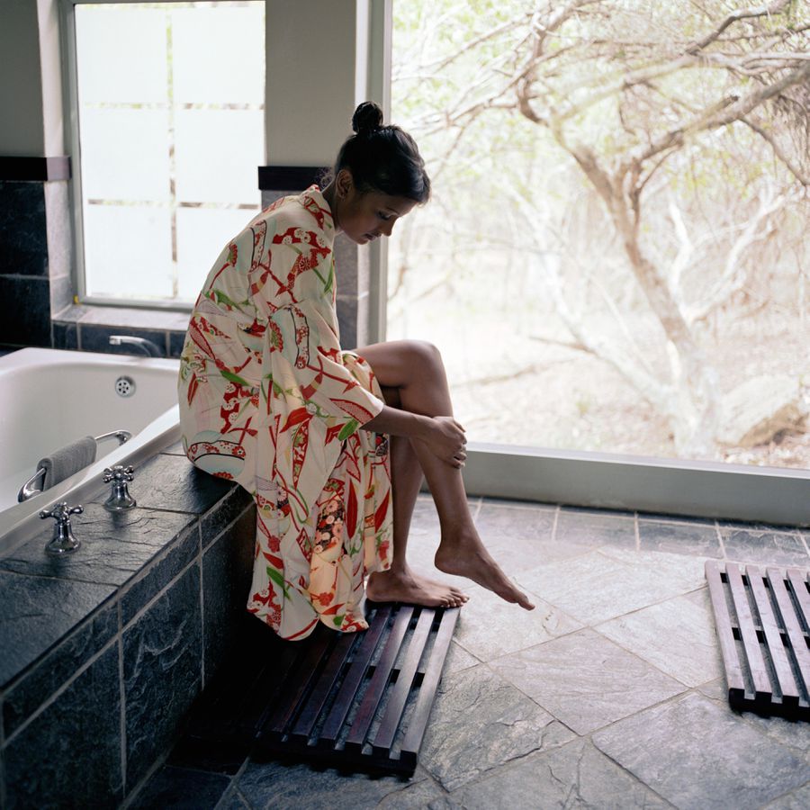 Person wearing a patterned robe sitting on the edge of a tub putting lotion on their legs 