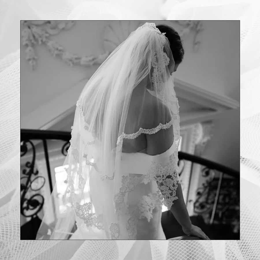 A black and white photo of a bride in a white wedding dress and a lace veil descending stairs on her wedding day.