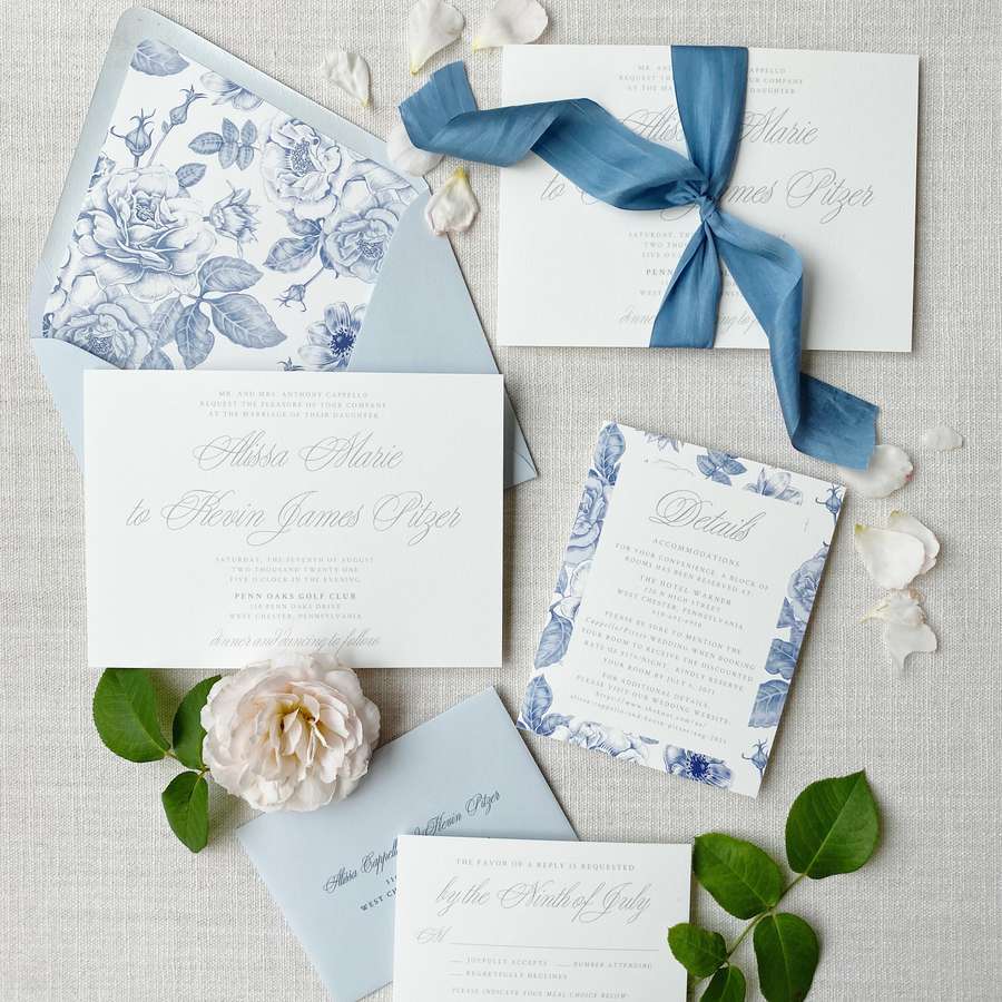 Blue and white wedding invitations with blue ribbon, white flower petals, and a floral envelope liner.