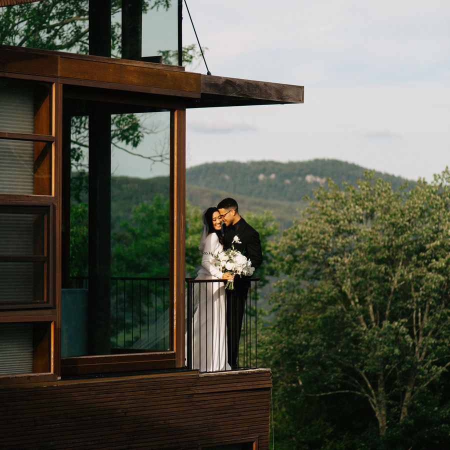 A bride and groom embracing on the balcony of an Airbnb wedding venue overlooking mountains and forrest. 