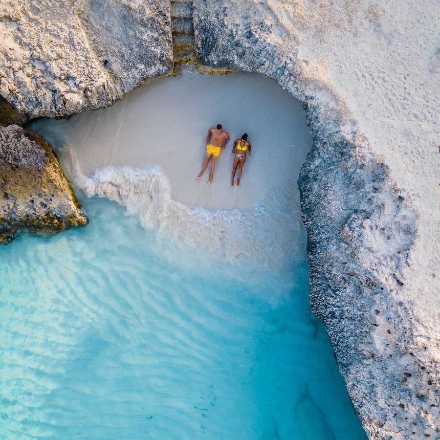 A couple in yellow bathing suits sitting on a private, white-sand beach in front of clear blue water in Aruba, a romantic honeymoon destination.