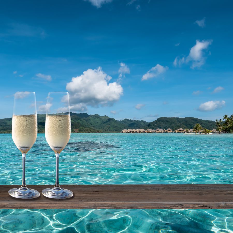 Two glasses of champagne served on the waterfront in Bora Bora, overlooking overwater bungalows and palm trees.