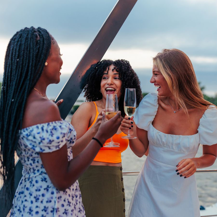 Woman in floral dress, woman in orange top and green skirt, and woman in white puff sleeve dress on a boat holding Champagne