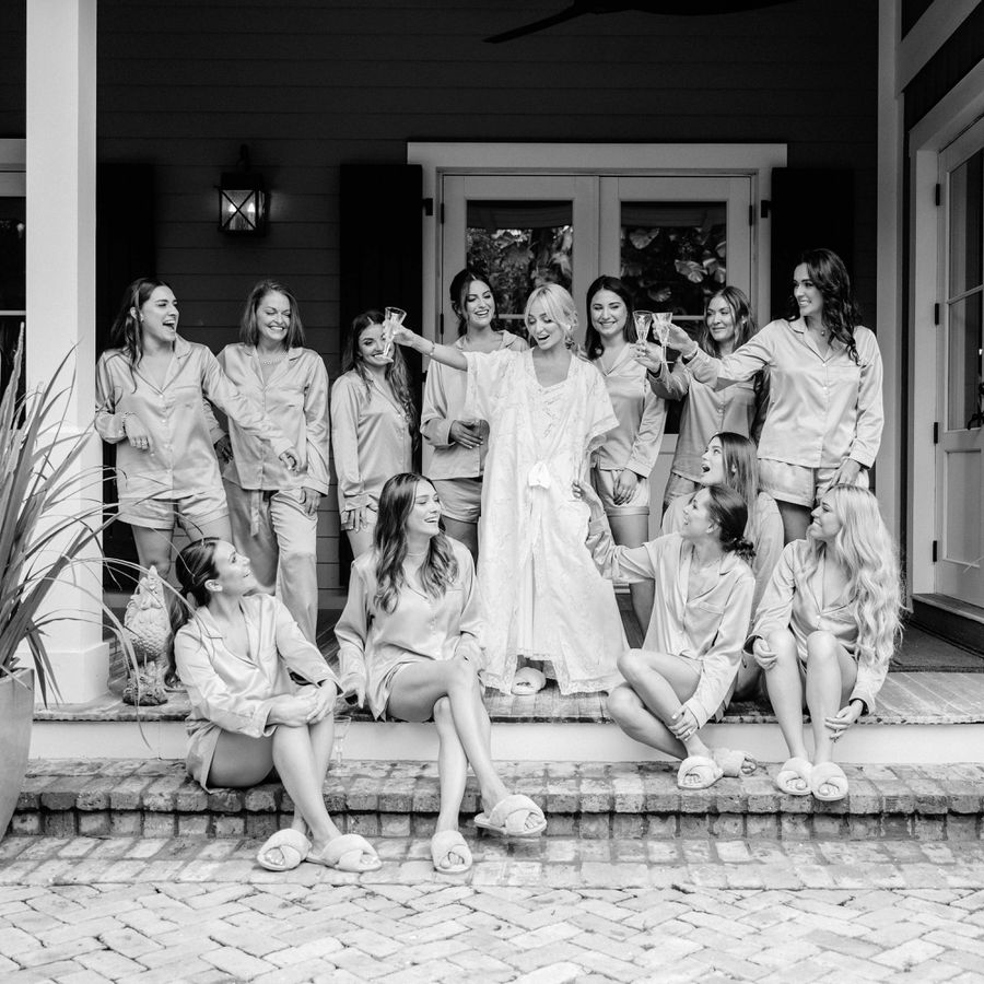Bride and Bridesmaids in Pajamas on Porch Raising Champagne Flutes in Cheers While Getting Ready for Wedding