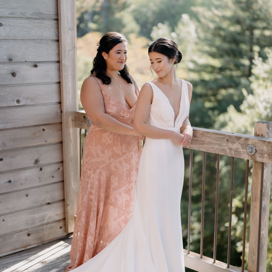 Bridesmaid with a half-up hairstyle in a peach dress helping the bride put on her wedding dress