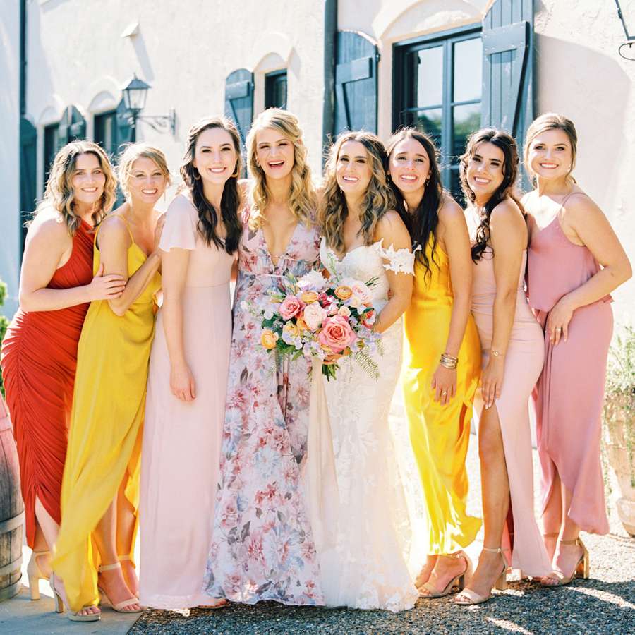 A bride in a white wedding gown with a rose bridal bouquet standing with bridesmaids and maids of honor in colorful dresses.