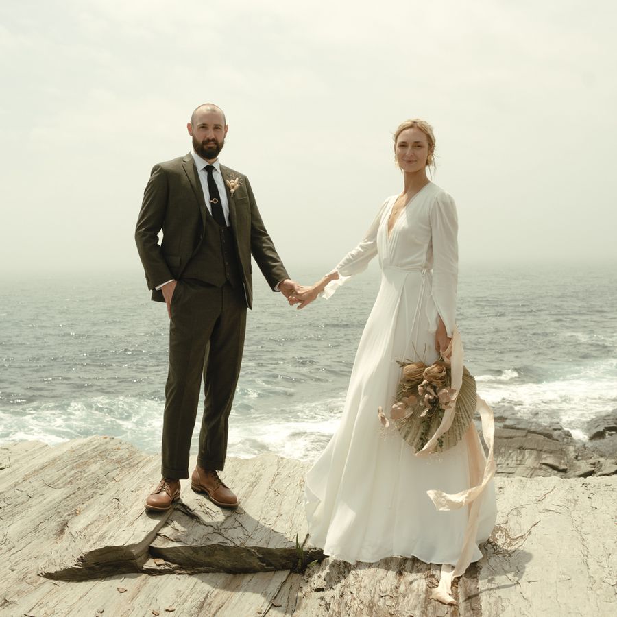 Groom in Brown Suit and Bride in Long Sleeve Reformation Wedding Dress Holding Hands on Cliffside in Maine