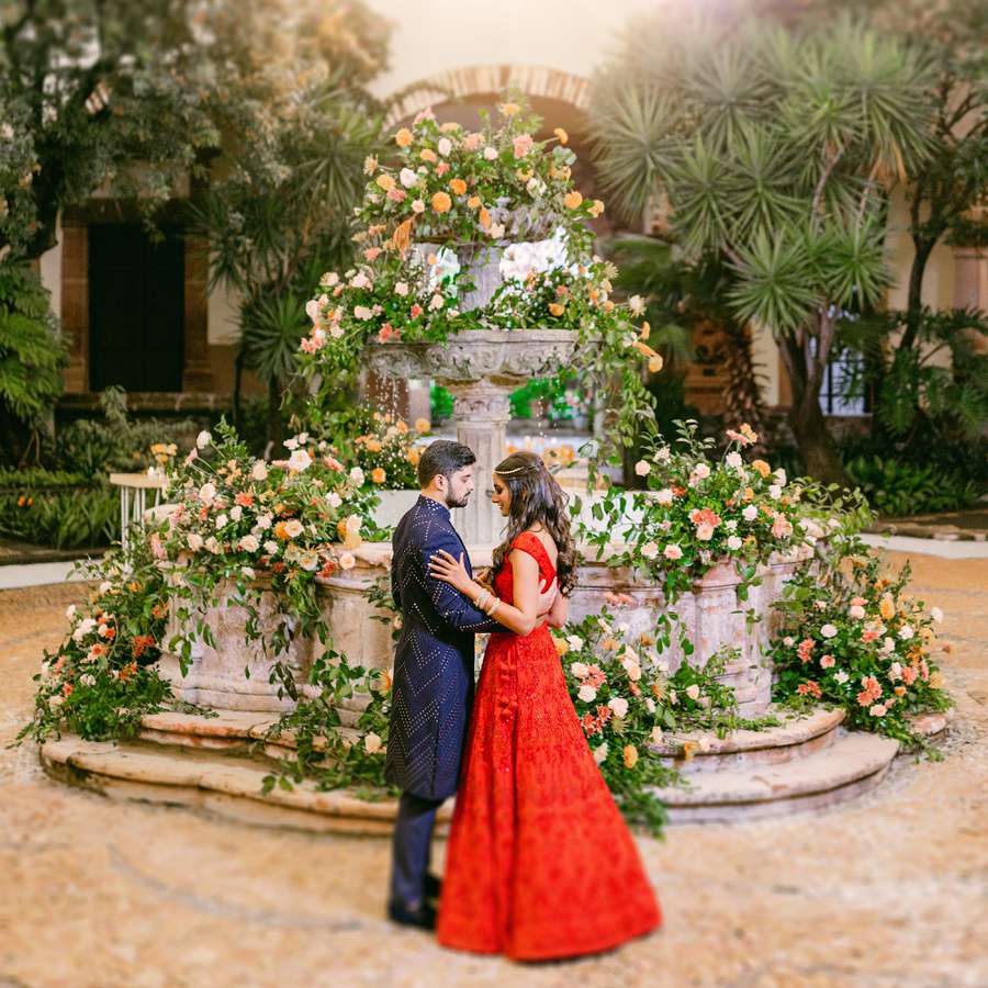 Groom in Navy Blue Sherwani and Bride in Red Lehenga Hugging in Front of Floral-Covered Fountain