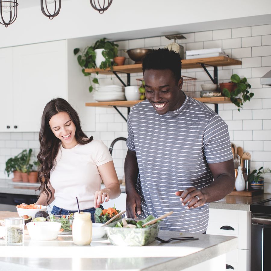 A happy couple cooks dinner together in a white-tiled kitchen decorated with plants.