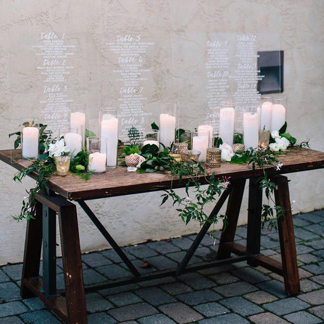 Wedding seating charts surrounded by candles and plants displayed on wood table on stone patio
