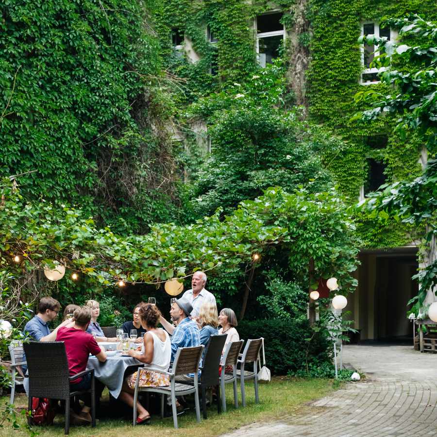 Family dinner outside of ivy-covered building in a courtyard with family members seated around table while father gives toast