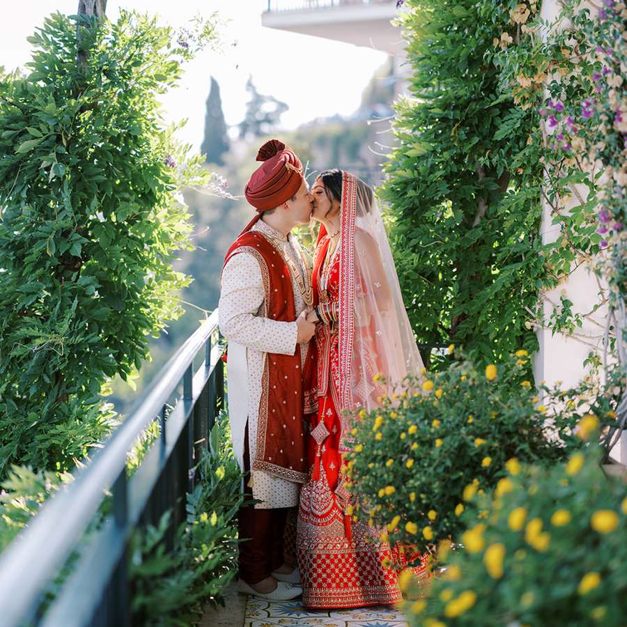 Bride and Groom in Traditional Indian Attire Kissing on Greenery-Covered Balcony in Amalfi, Italy