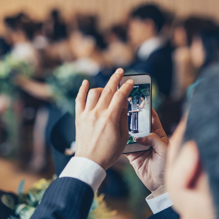 A wedding guest taking a photo on an iPhone of a bride and groom getting married at a wedding ceremony.
