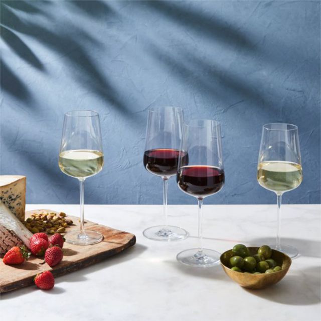 Crystal glassware full of red and white wine displayed on a white surface next to a grapes, cheese, and strawberries