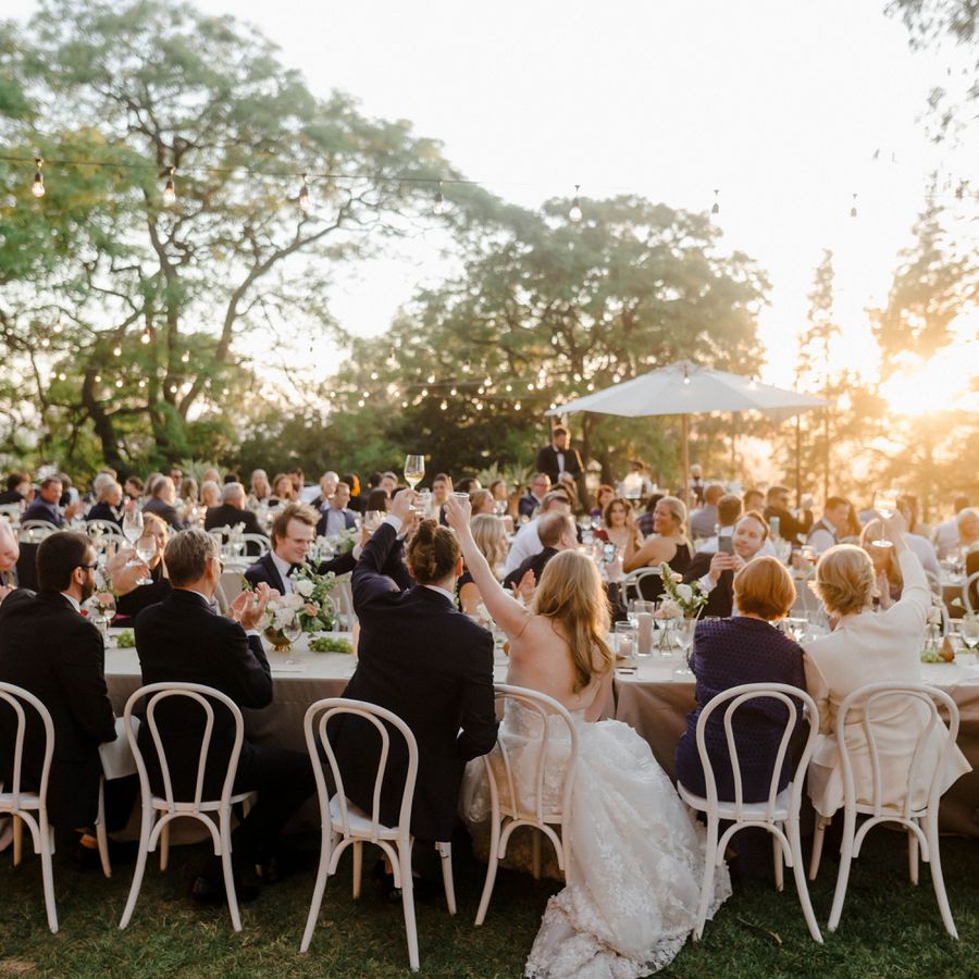 bride, groom, and wedding guests raising wine glasses while seated at tables during sunset wedding reception