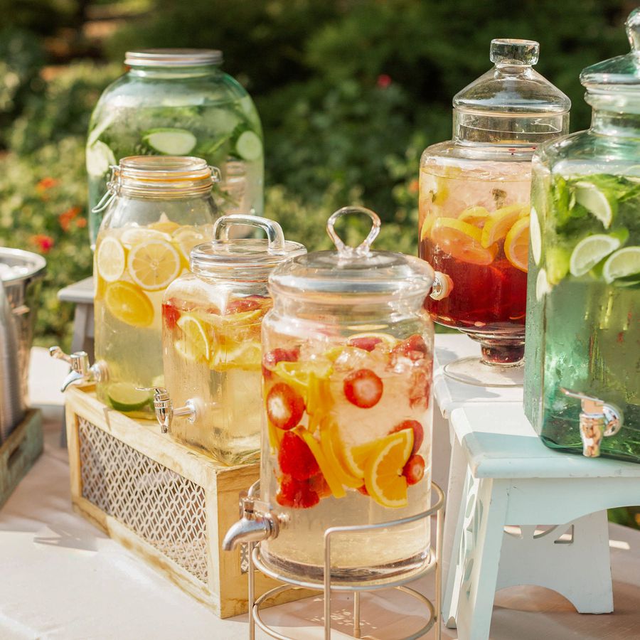 Five glass dispensers with infused water featuring strawberries, oranges, lemons, limes, cucumbers, and mint
