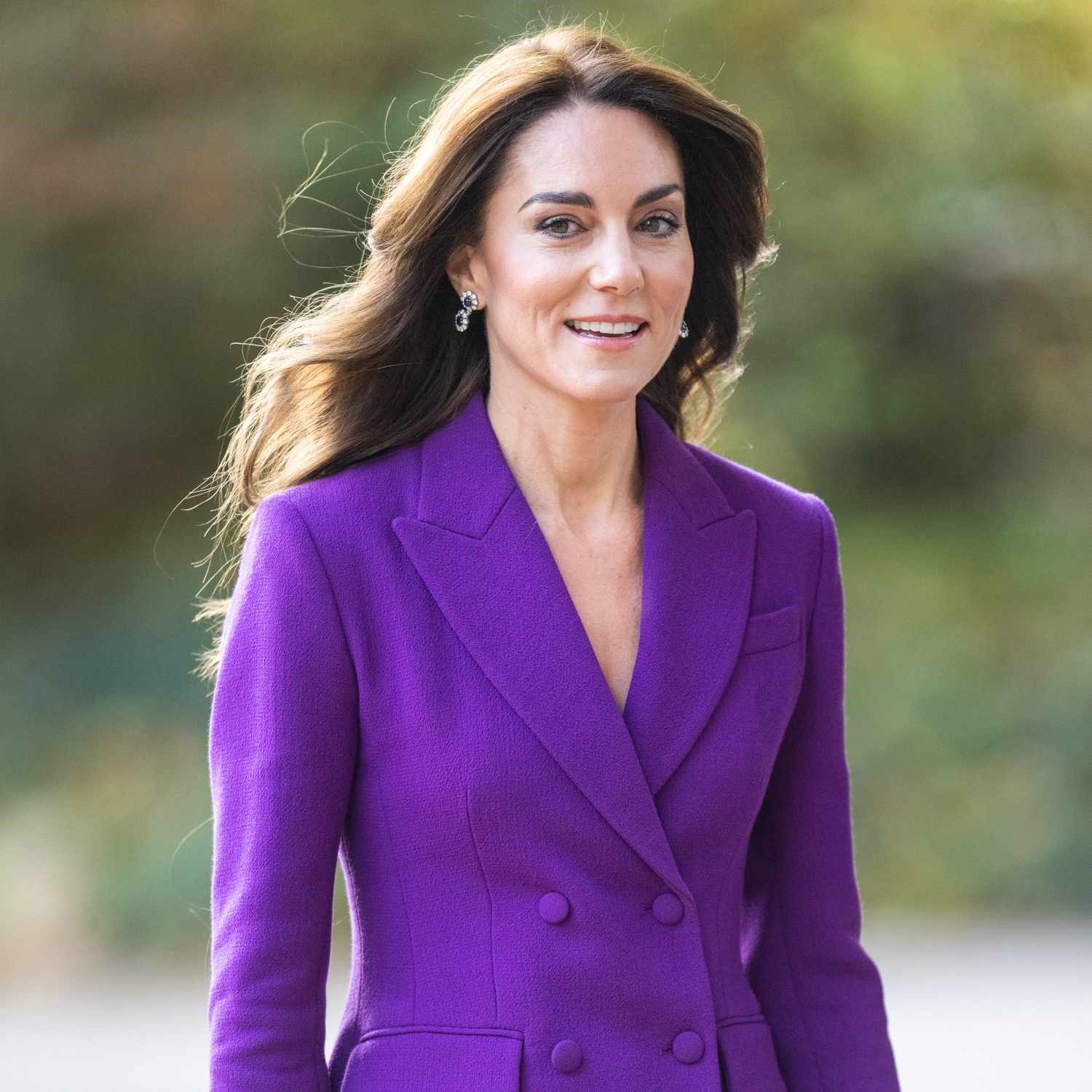 Kate Middleton smiling in a purple suit with her hair blowing in the wind
