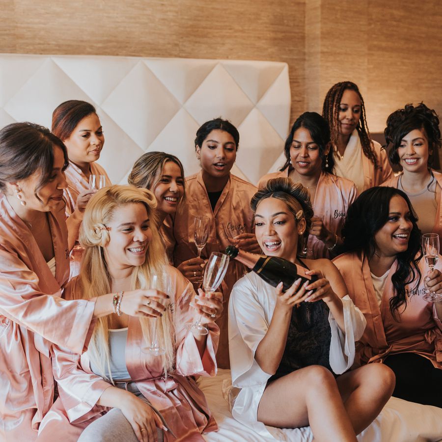 A bride pouring pink Champagne sitting on a bed in a hotel room with her bridesmaids who are wearing pink silk monogrammed robes and smiling.