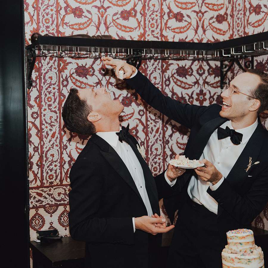 Same-sex couple feeding each other wedding cake in an intimate indoor reception