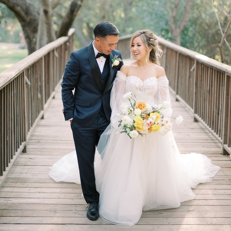Portrait of Groom in Navy Suit Walking with Bride in Off-the-Shoulder Wedding Dress Carrying White and Yellow Bouquet