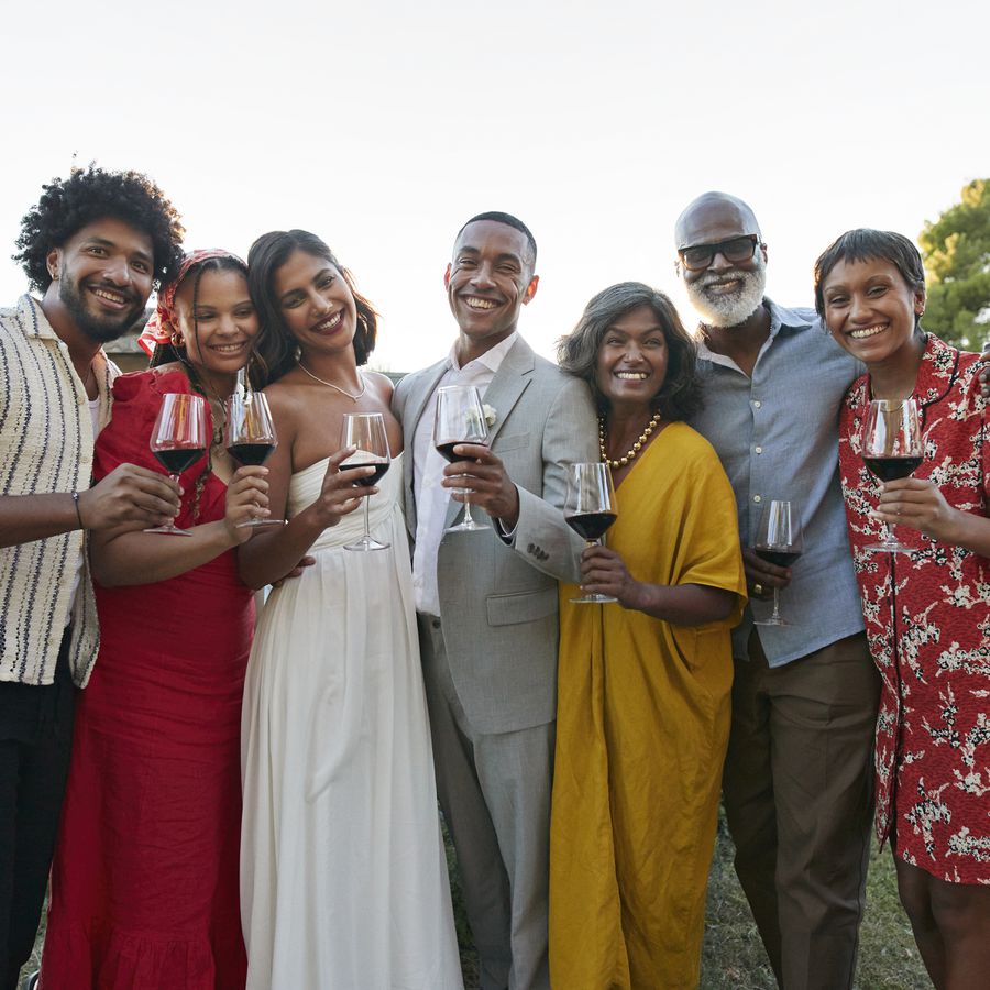 A bride and groom smiling with their adult wedding guests and holding red wine at their reception