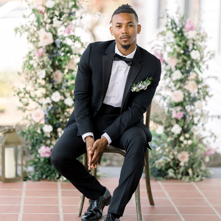 groom sitting on a chair while posing for a wedding portrait in front of a floral background, wearing a black tuxedo and a bowtie