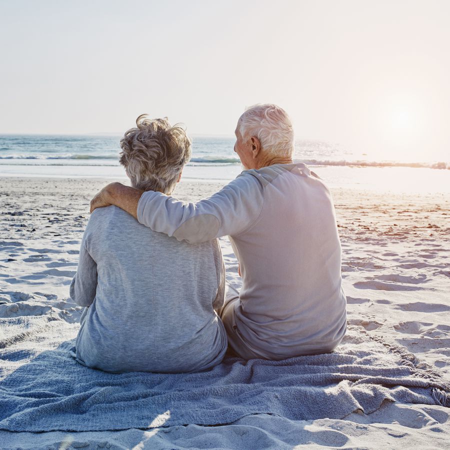 An elderly husband and wife sit on the beach together during sunset, with the husband's arm over his wife's shoulders.