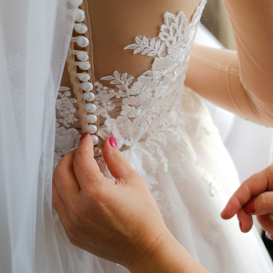 A bridesmaid wearing pink nail polish helps the bride button up her white wedding gown.