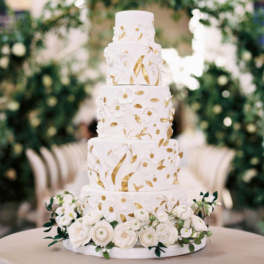 Four-tiered white wedding cake with sugar flowers and gold leaves with white florals at base