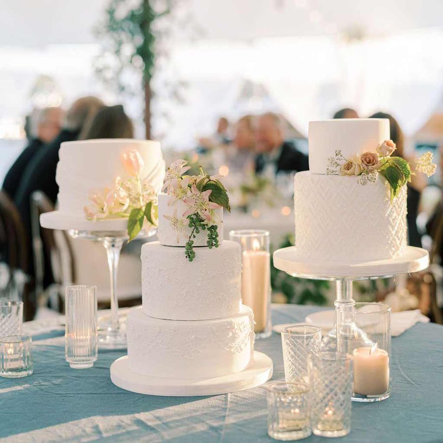 Three white lace wedding cakes of varying size on cake stands decorated with pink and green florals