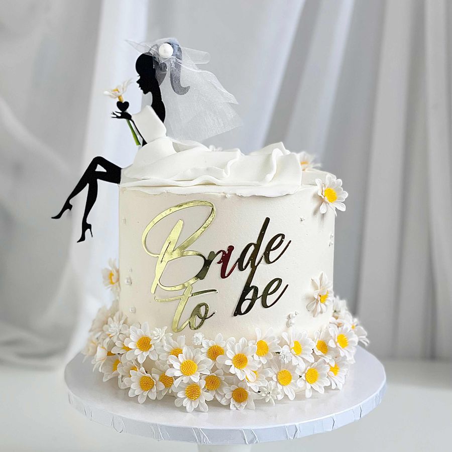 One-tier white cake with daisies, a "Bride to be" gold sign, and a silhouette of a bride in a veil with white fondant ruffles
