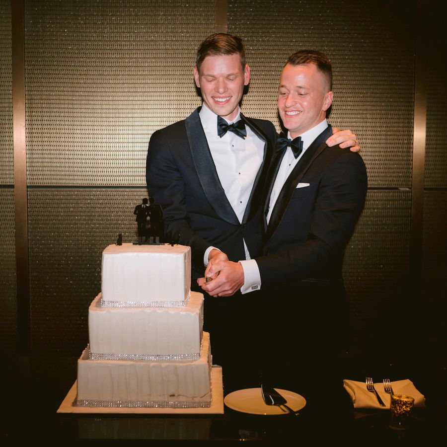 Two grooms in tuxedos cut into a white three-tier wedding cake with a black silhouette cake topper of grooms kissing
