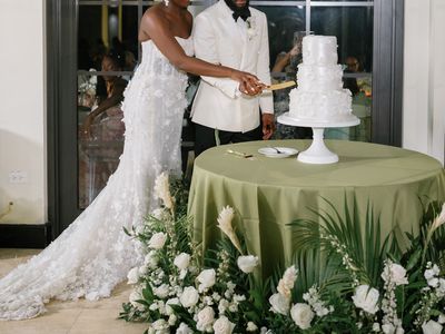 Bride in white wedding gown cutting three-tier wedding cake with groom wearing a white formal jacket, surrounded by white flowers.