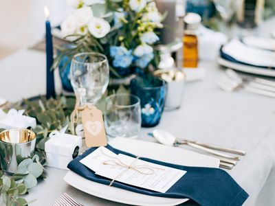 A navy blue napkin and a white menu tied together with a string on top of a white plate set on a table with white linens, greenery, white flowers, and blue taper candles