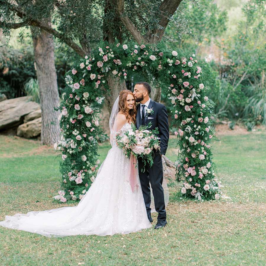 Portrait of Bride in Strapless Wedding Dress and Groom in Black Tuxedo in Front of Floral Arch