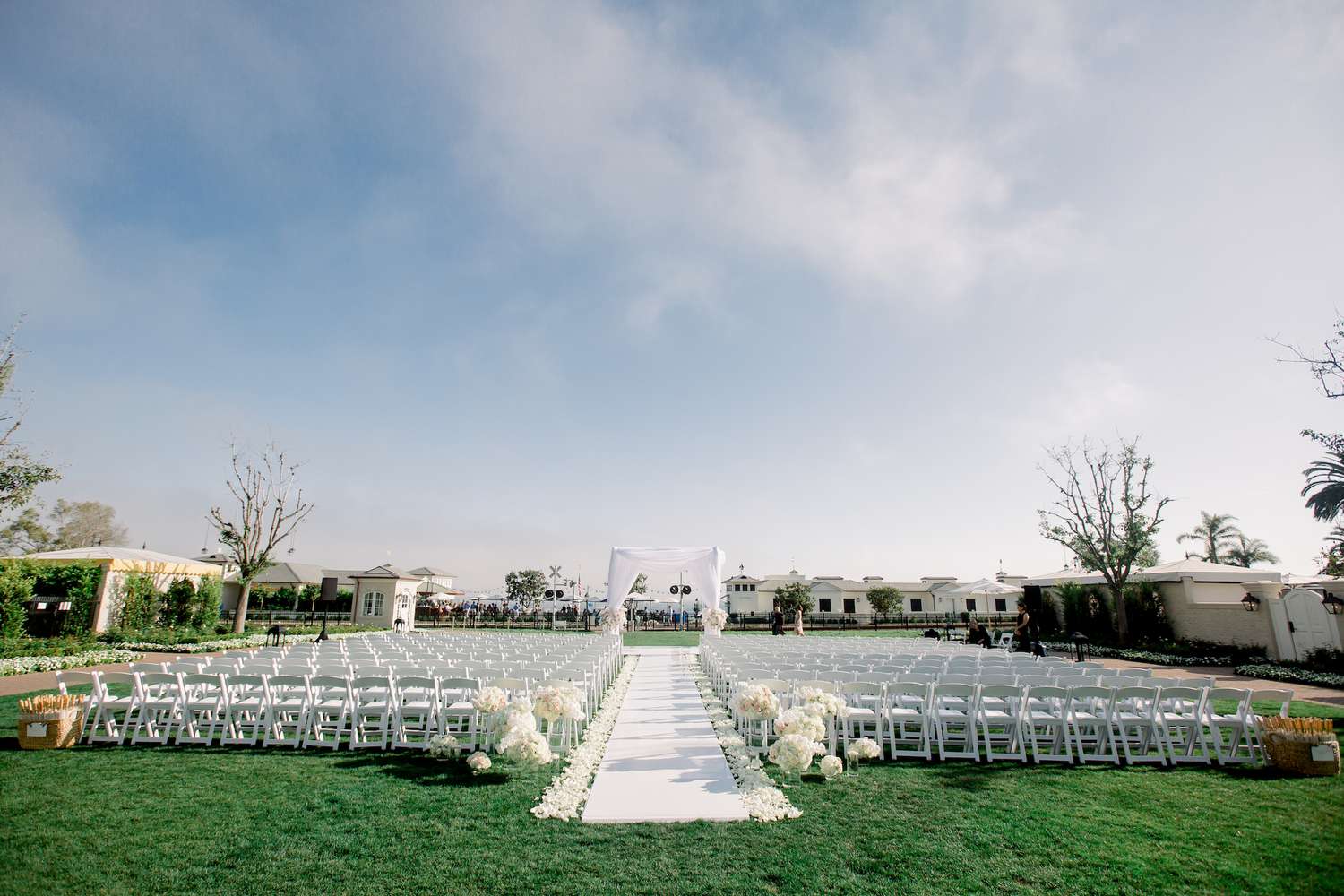 Backyard ceremony with white runner, white petals lining the aisle, and varying heights of glass vases filled with white flowers at the start