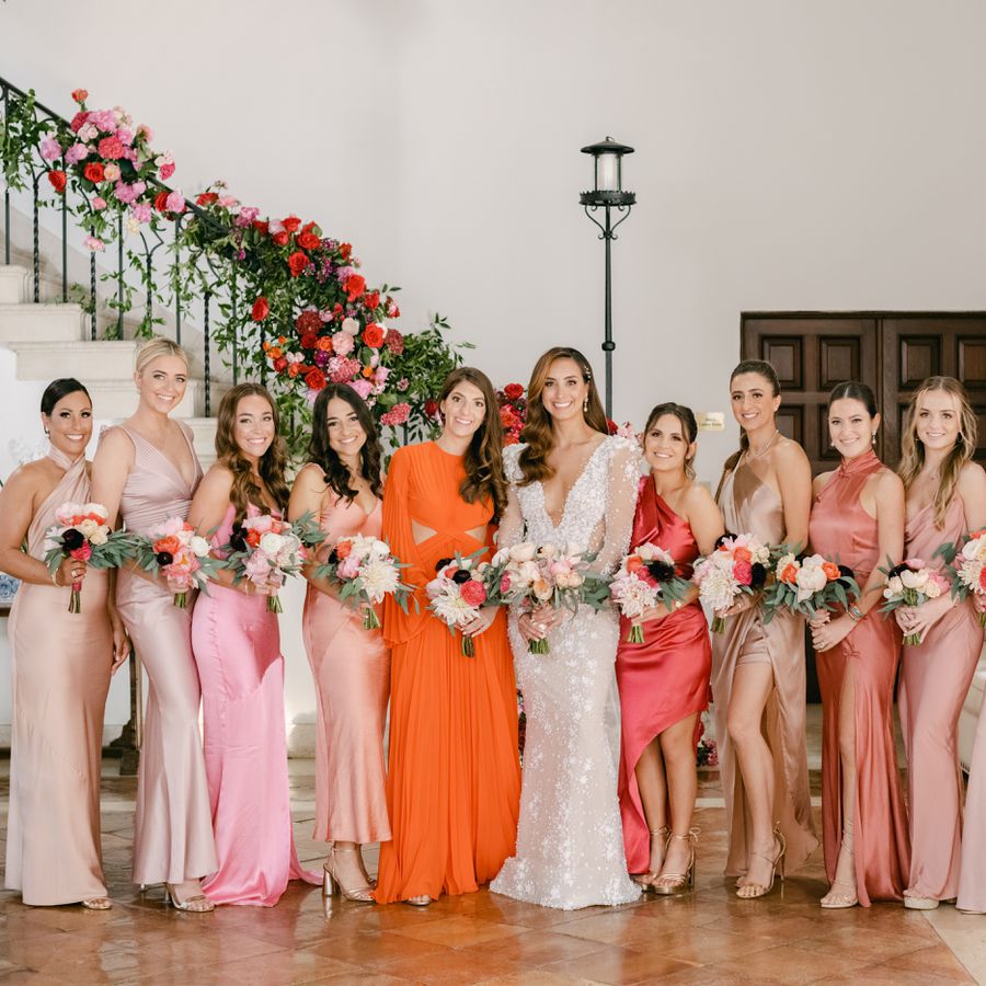 Bride posing with bridal party bridesmaids wearing assorted dresses in front of staircase with flowers on railing