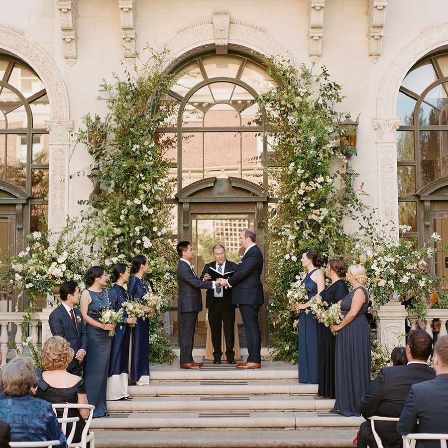 Grooms Holding Hands at Outdoor Wedding Ceremony with Greenery and Floral Backdrop