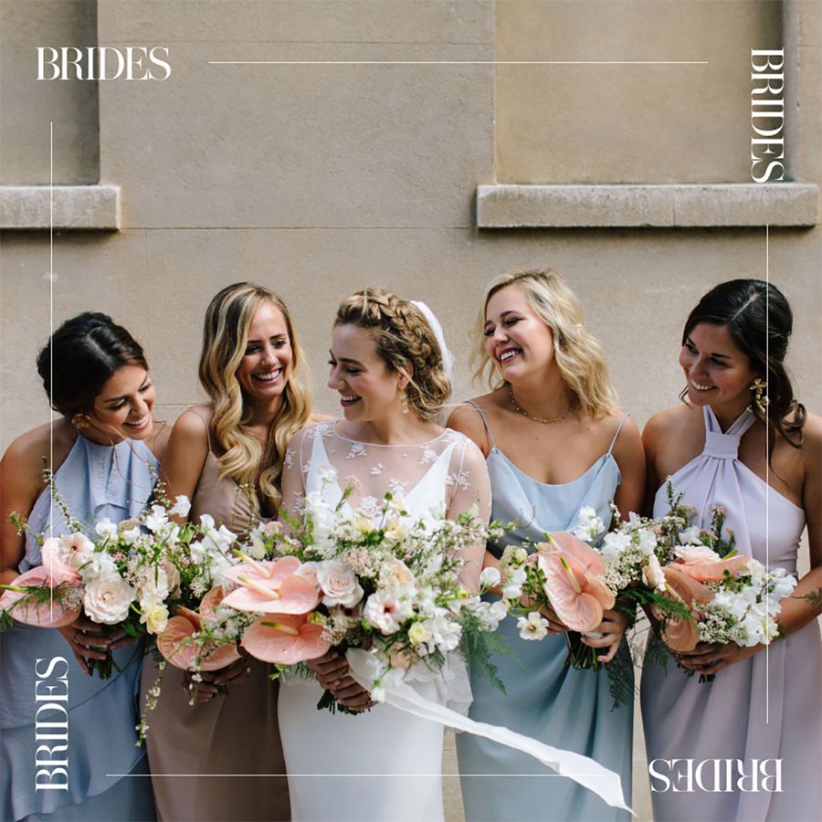 Bride surrounded by bridesmaids wearing pastel dresses while holding flowers and standing in front of a beige building
