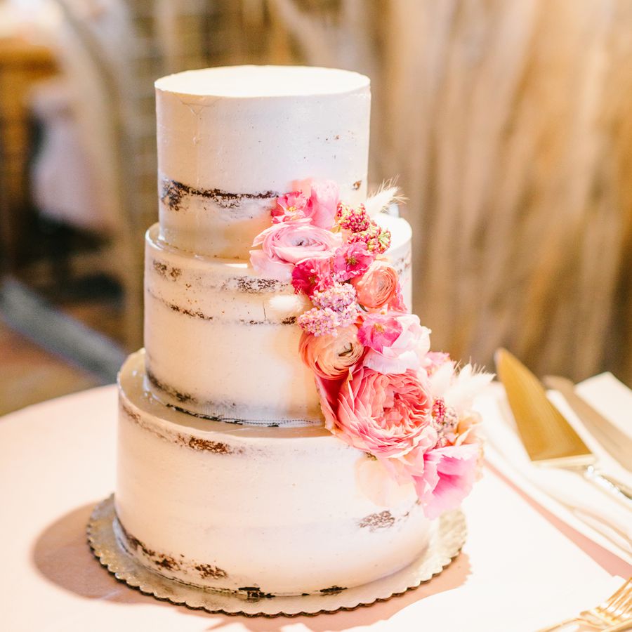 White three-tiered semi-naked wedding cake with cascading fresh pink flowers served at wedding reception.