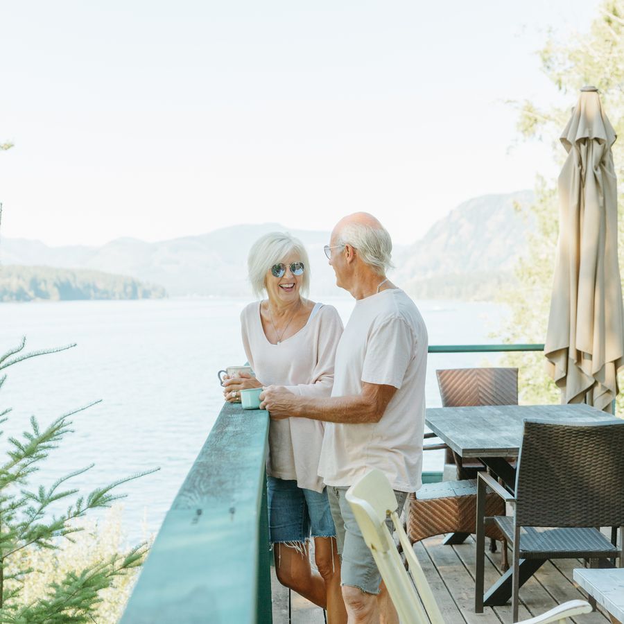 Older couple standing on a deck with cups of coffee, with a lake and mountains in the background 