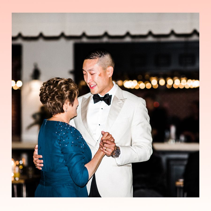 A groom smiling at his mother, who is wearing a blue dress, as they do a mother-son dance during a wedding reception.